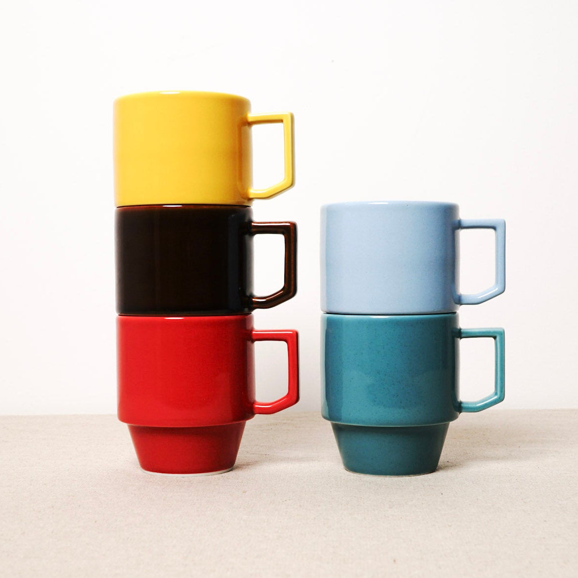 Stacked ceramic mugs in yellow, black, red, and blue on a light background