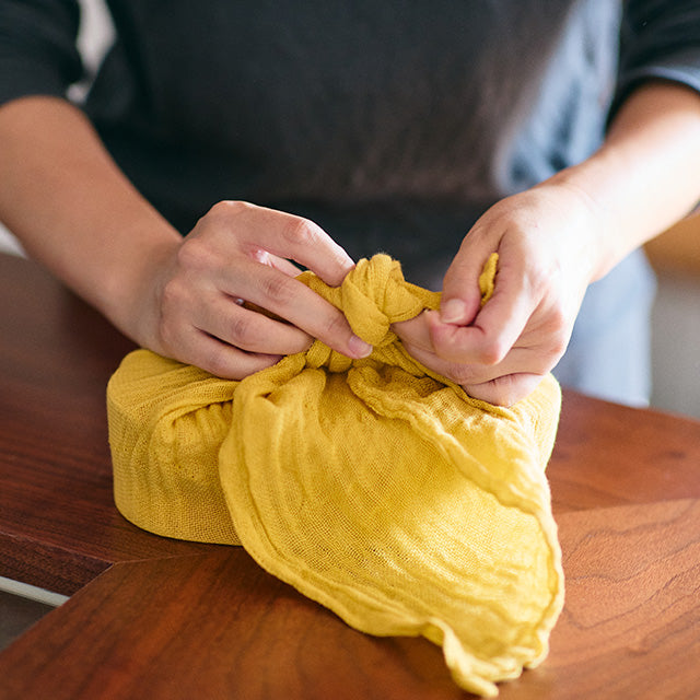 Person wrapping a lunch box with yellow fabric on a wooden table