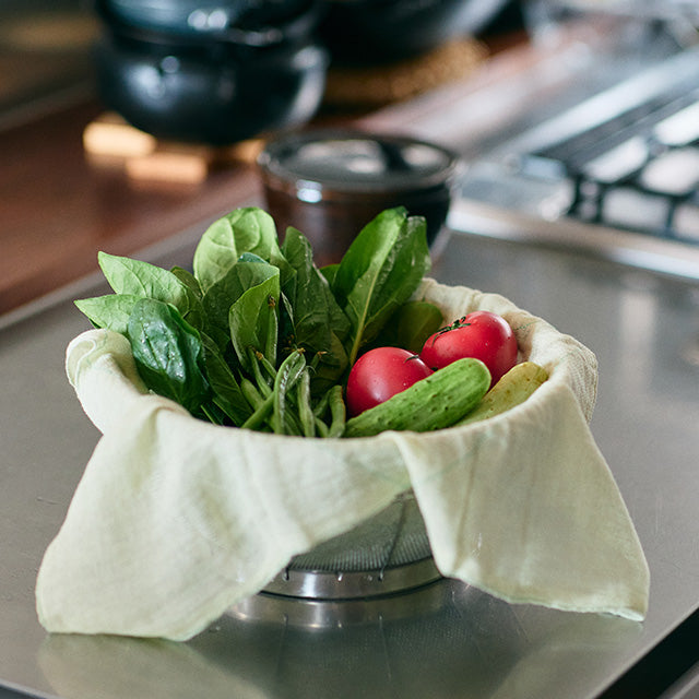 Fruit and vegetables covered in a white tea towel in a strainer on a kitchen counter