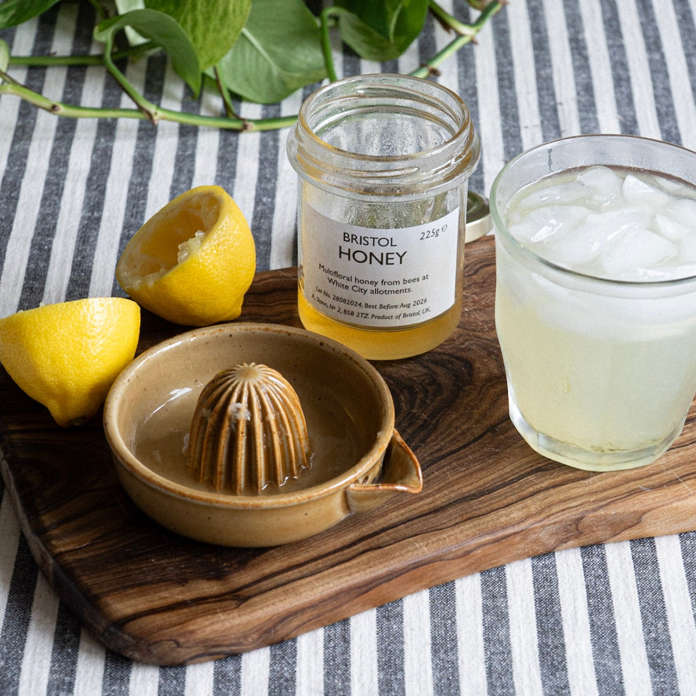 Ceramic lemon squeezer, Jar of Bristol Honey, lemon, and a glass of lemonade on a striped tablecloth.