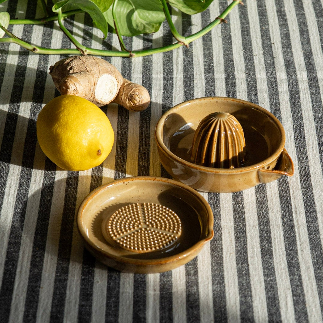 Ceramic grater and lemon squuzer on a striped tablecloth with a lemon and ginger root.