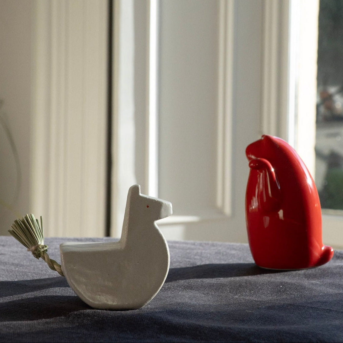 Two ceramic birds, one white and one red, on a surface with a window in the background.