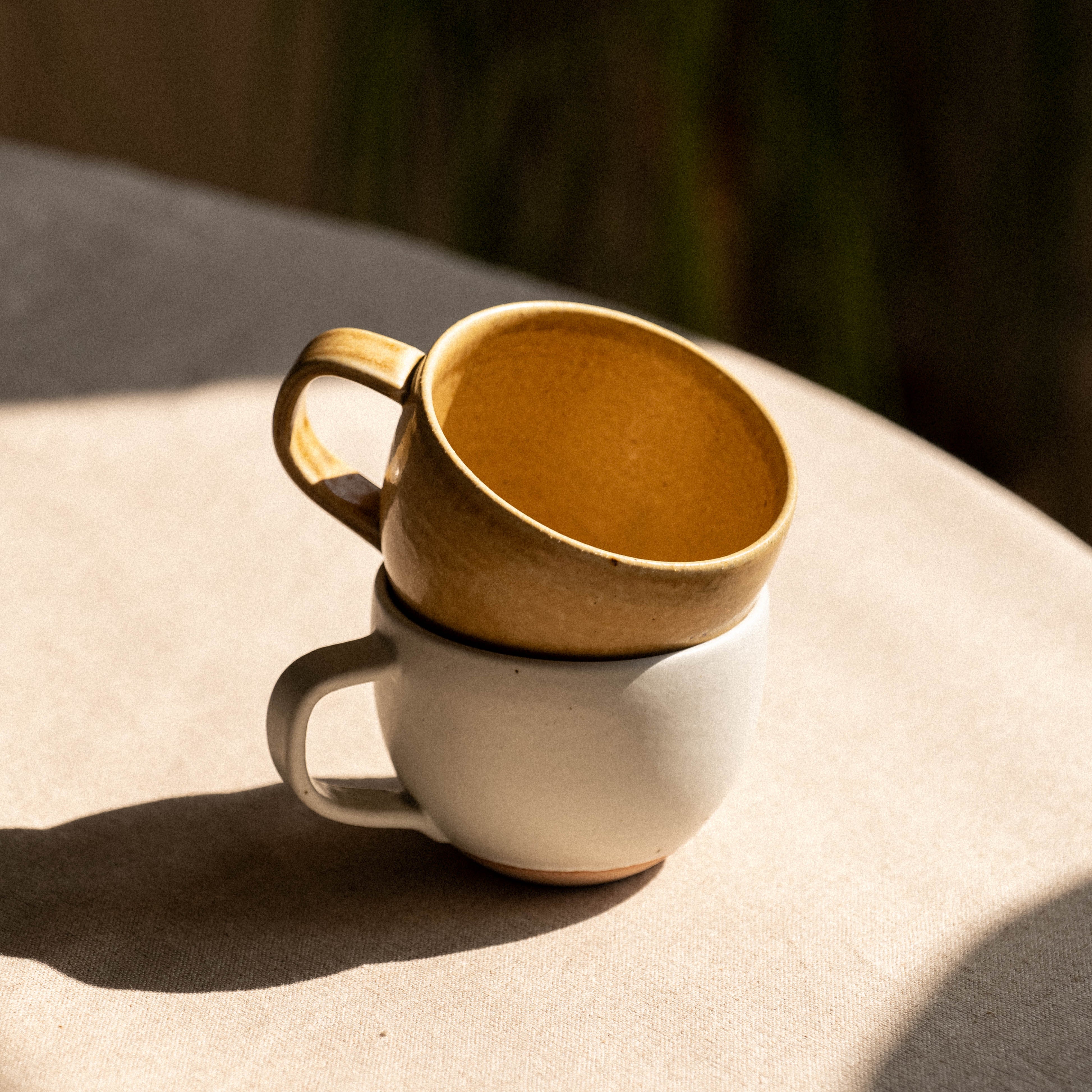 Two ceramic cups, one white and one mustard yellow, stacked on a light surface with a blurred natural background.