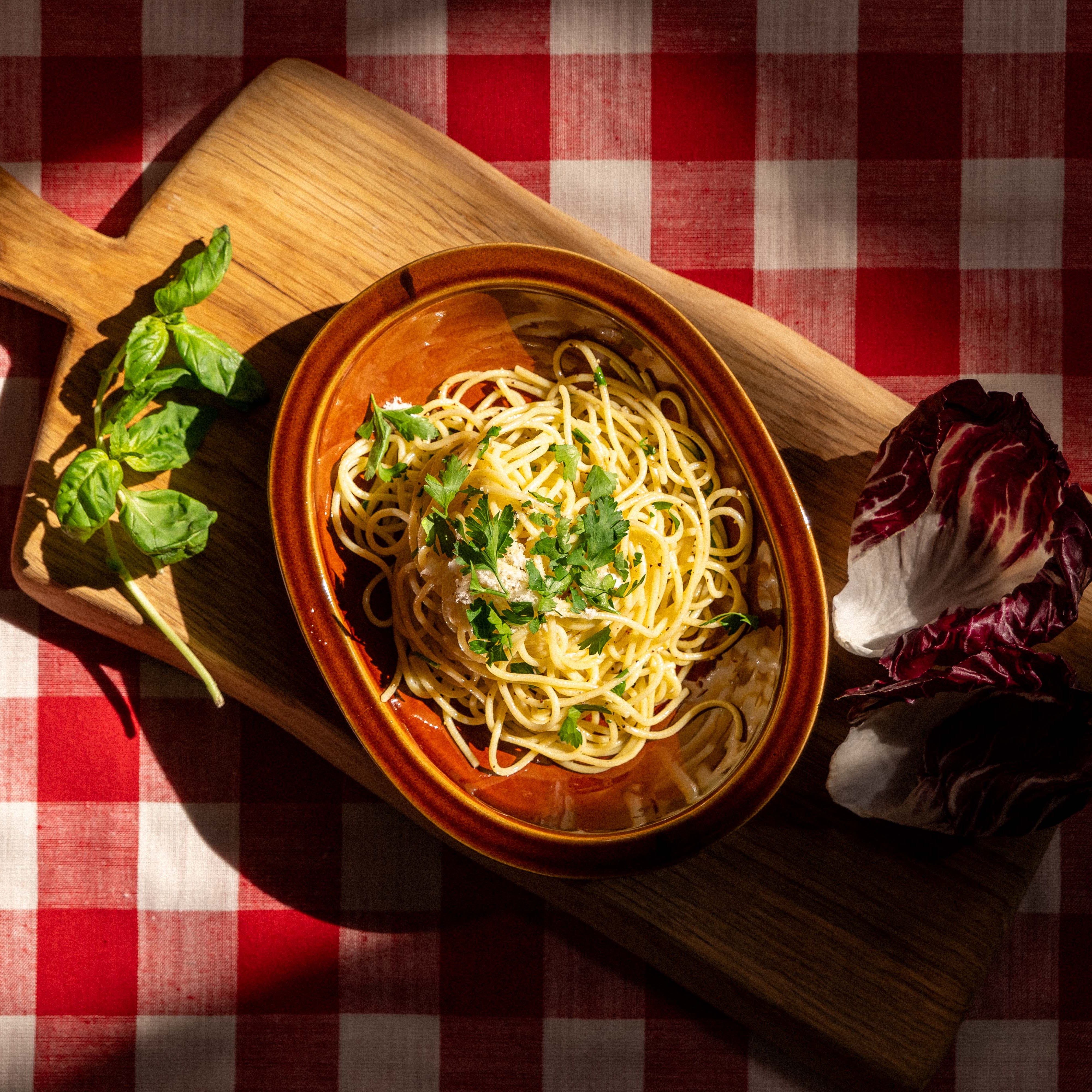 Bowl of pasta with greens on a wooden board over a red and white checkered tablecloth