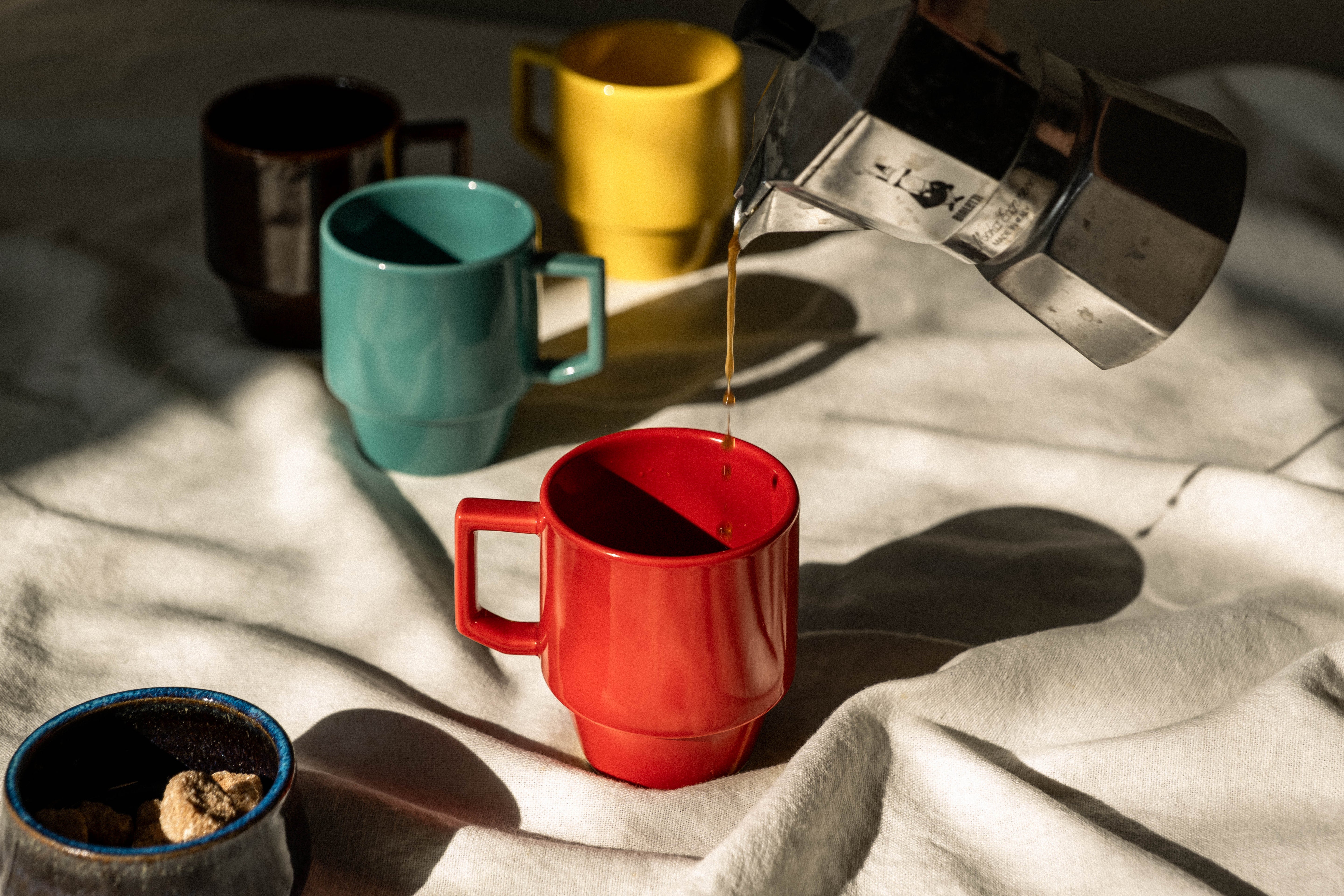 Coffee being poured from a kettle into colorful mugs on a textured surface.