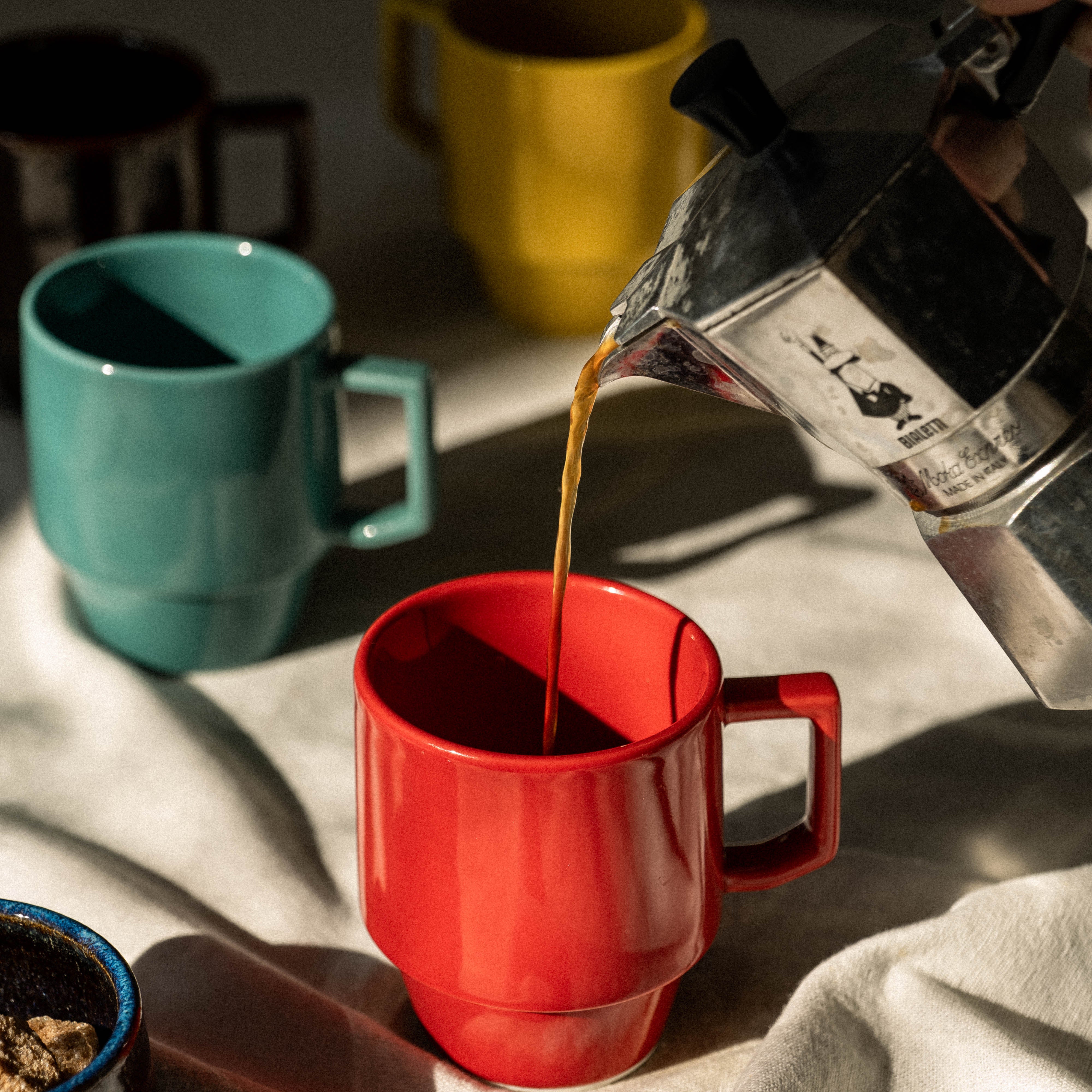 Coffee being poured from a French press into a red mug with a yellow mug and blue cup in the background.