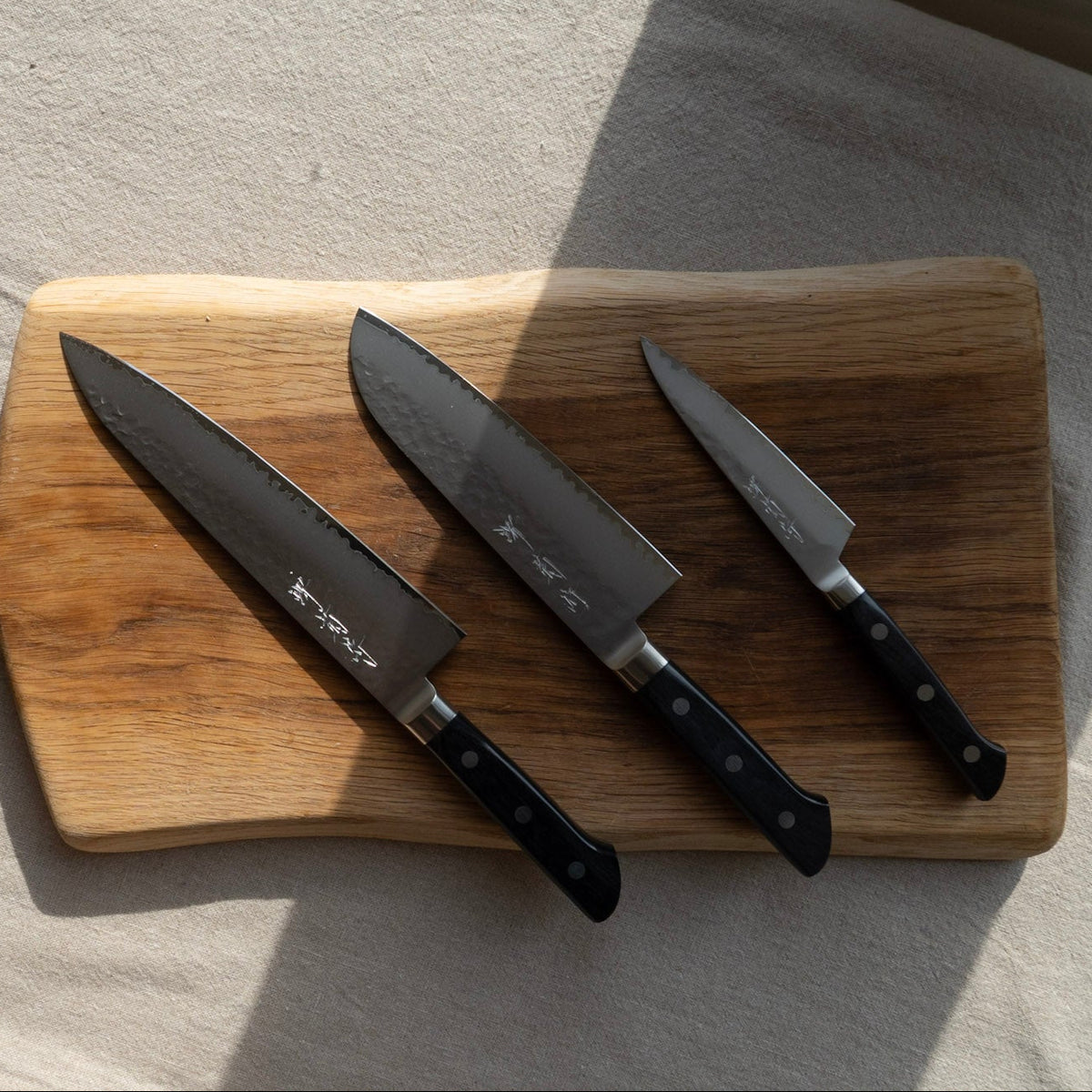 Three Japanese Sakai kitchen knives with black handles and textured steel blades are arranged on a wooden cutting board.