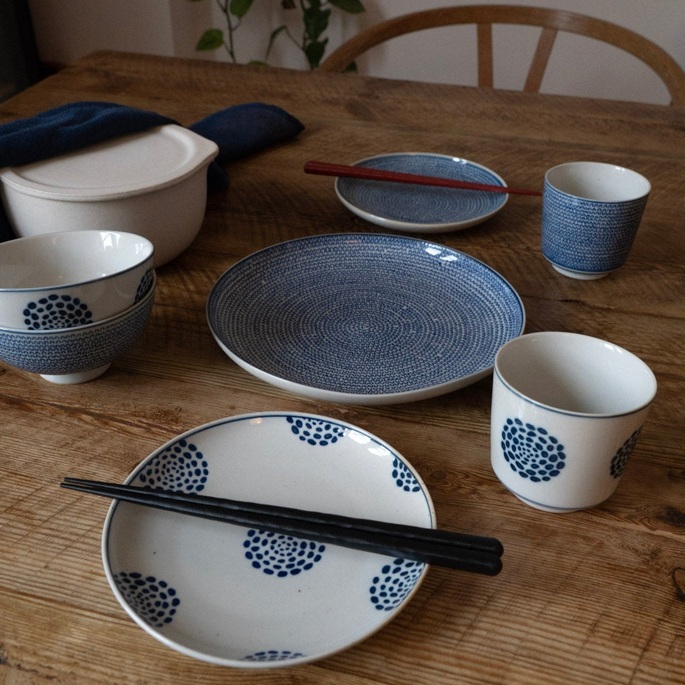 Ceramic tableware including bowls, plates, and cups on a wooden table with a plant in the background.