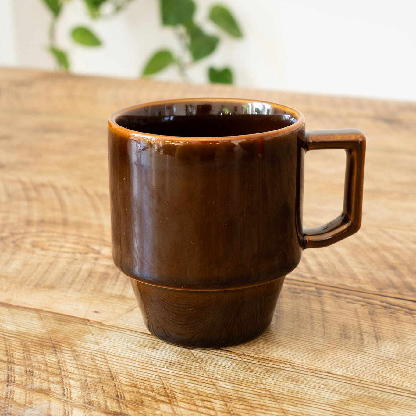 Brown ceramic mug on a wooden surface with a blurred green plant in the background