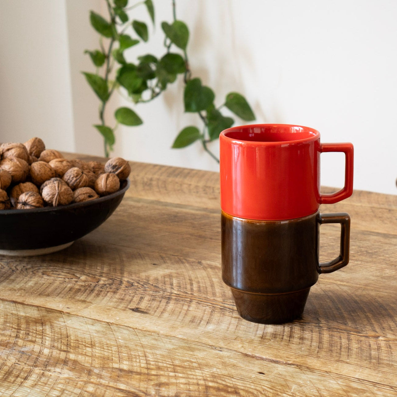 Red and brown ceramic mug on a wooden table with a bowl of nuts and a plant in the background.