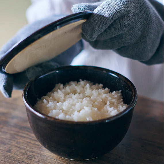 Person wearing gloves opening a black container to reveal rice in a bowl on a wooden surface.
