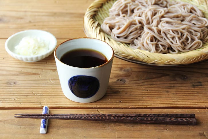 Soba noodles with a cup of soup, chopsticks, and a small bowl on a wooden surface.