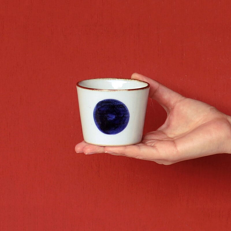 Hand holding a small white cup with a blue circular design against a red background