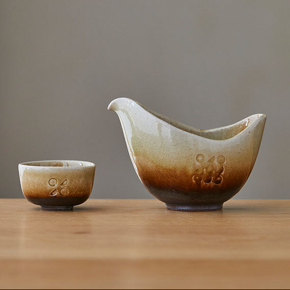 A ceramic sake pitcher and matching small sake cup with a brown and beige gradient glaze, displayed on a wooden surface against a neutral gray background.