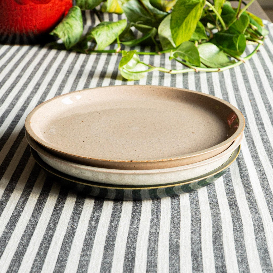 Three ceramic plates stacked on a striped tablecloth with a red vase and green plant in the background.