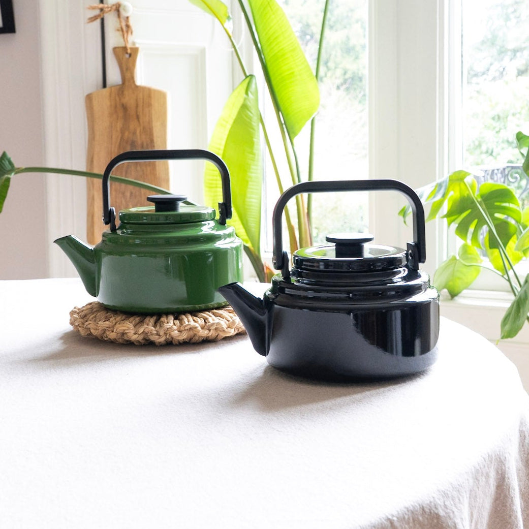 Two teapots, one green and one black, on a table with plants in the background.