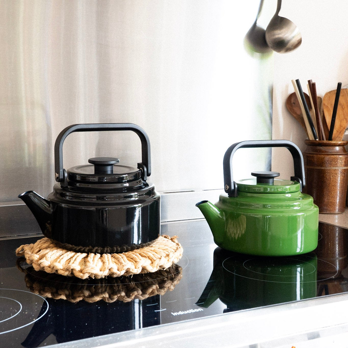 Two teapots, one black and one green, on a stovetop with kitchen utensils in the background.