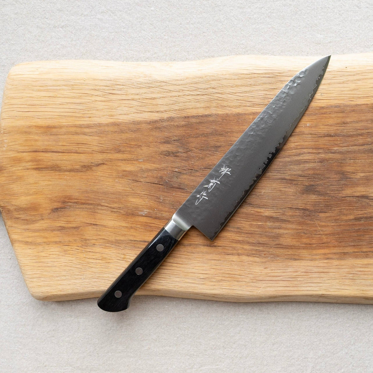 A Japanese Sakai kitchen knife with a dark textured blade and black handle, resting diagonally on a rectangular wooden cutting board.