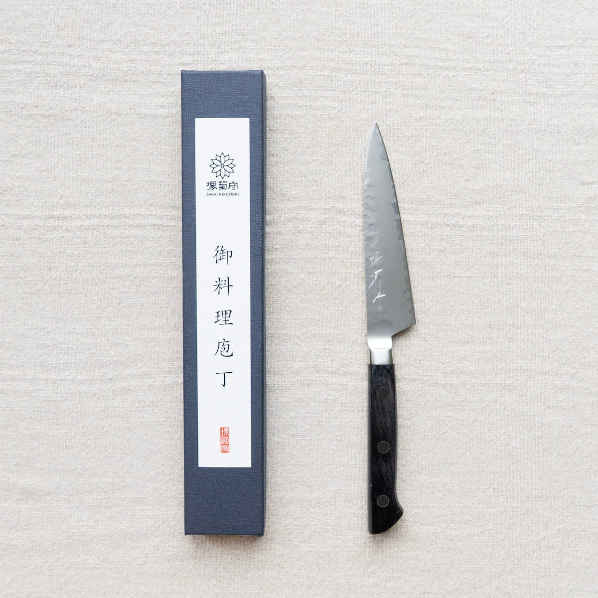 A Japanese Sakai kitchen knife with a black handle and silver blade, displayed next to its dark blue  box. 
