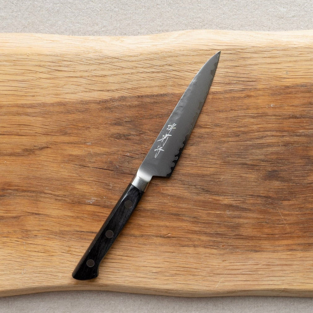 A Japanese-style Sakai kitchen petty knife with a dark textured steel blade and a black handle, resting diagonally on a rustic live-edge wooden cutting board.