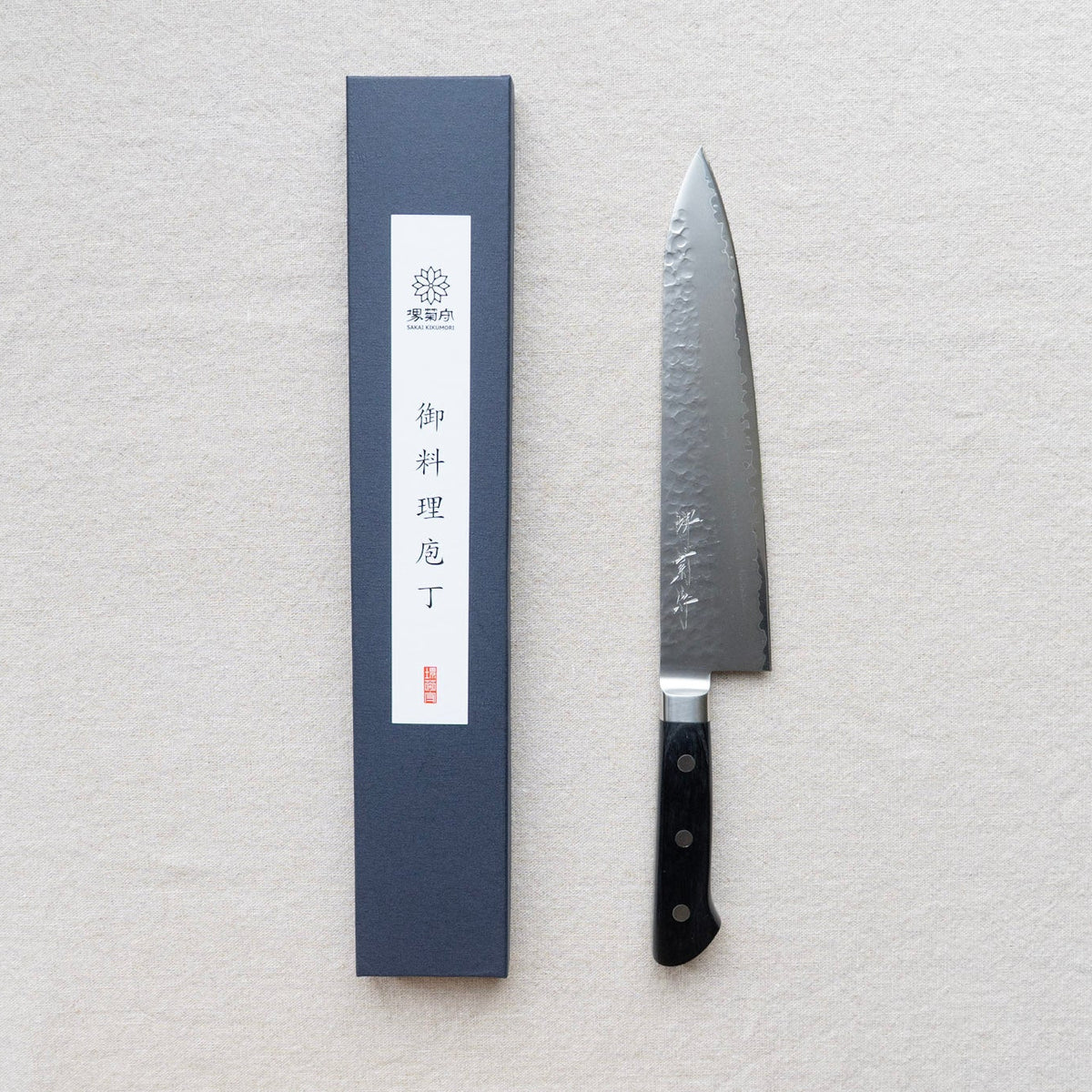 A Japanese kitchen knife with a hammered blade and black handle, displayed next to its dark blue presentation box on a textured off-white fabric background.