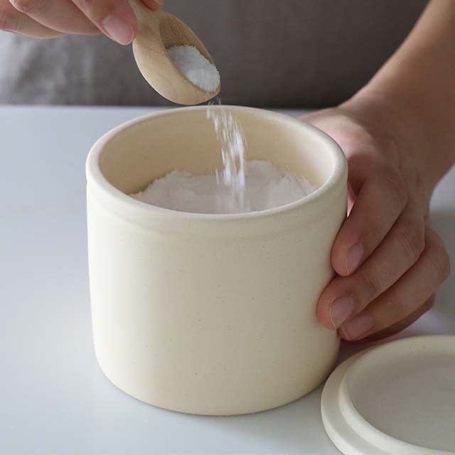 Person adding salt to a ceramic container with a wooden spoon.